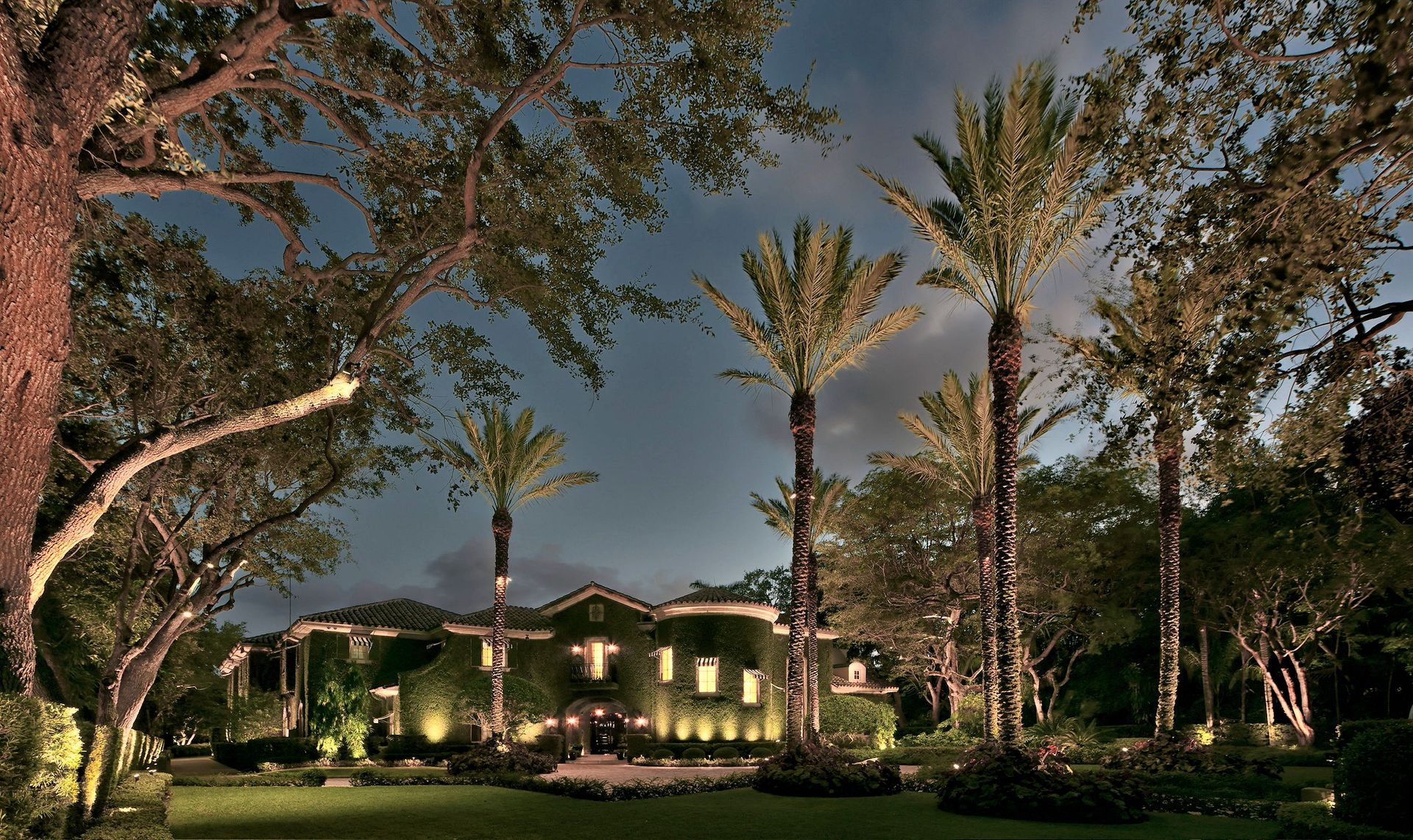 A sprawling, vine-covered Mediterranean estate illuminated at dusk, surrounded by tall palm trees and lush landscaping.