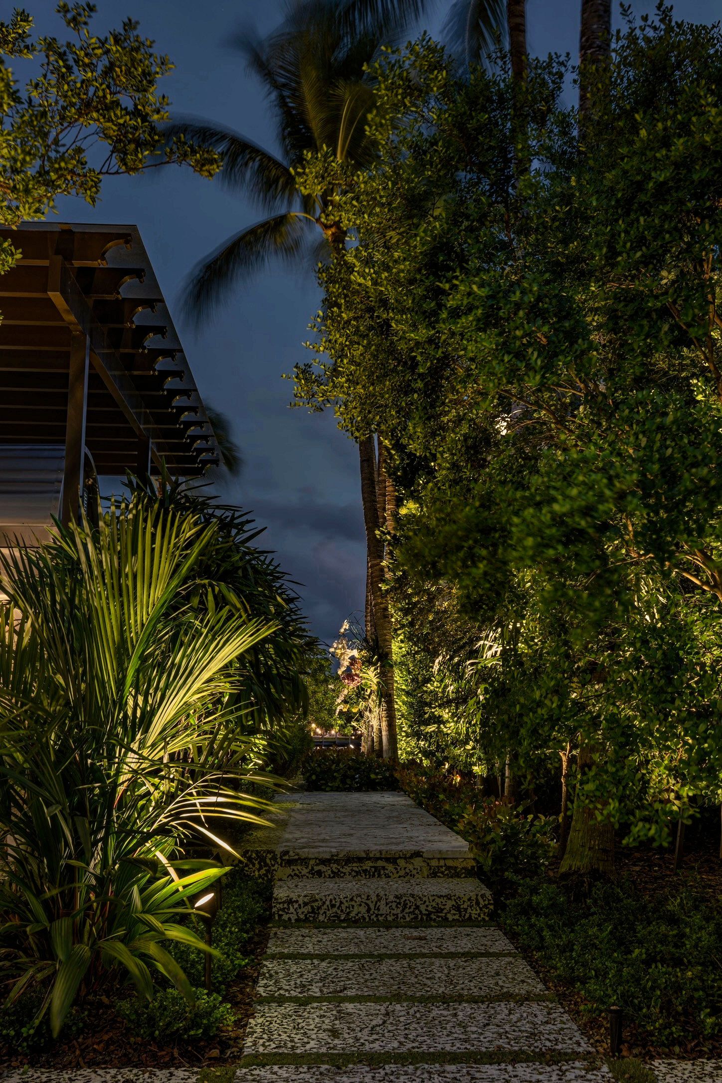 A stone walkway illuminated by garden lights leads through lush green trees and palms under a twilight sky.