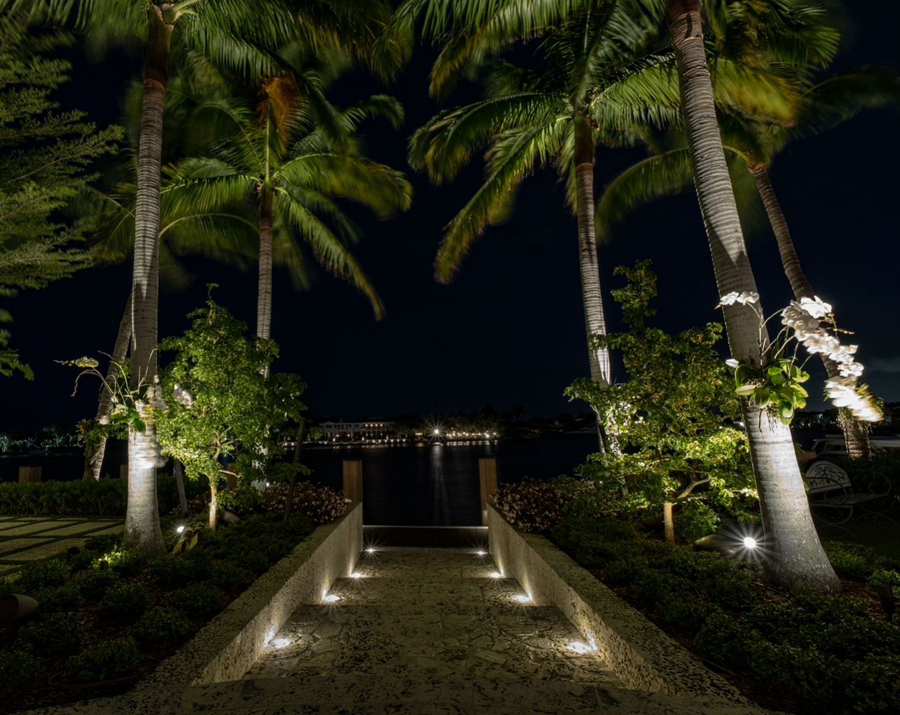 A lit stone pathway leads through palm trees toward a dark waterfront at night.