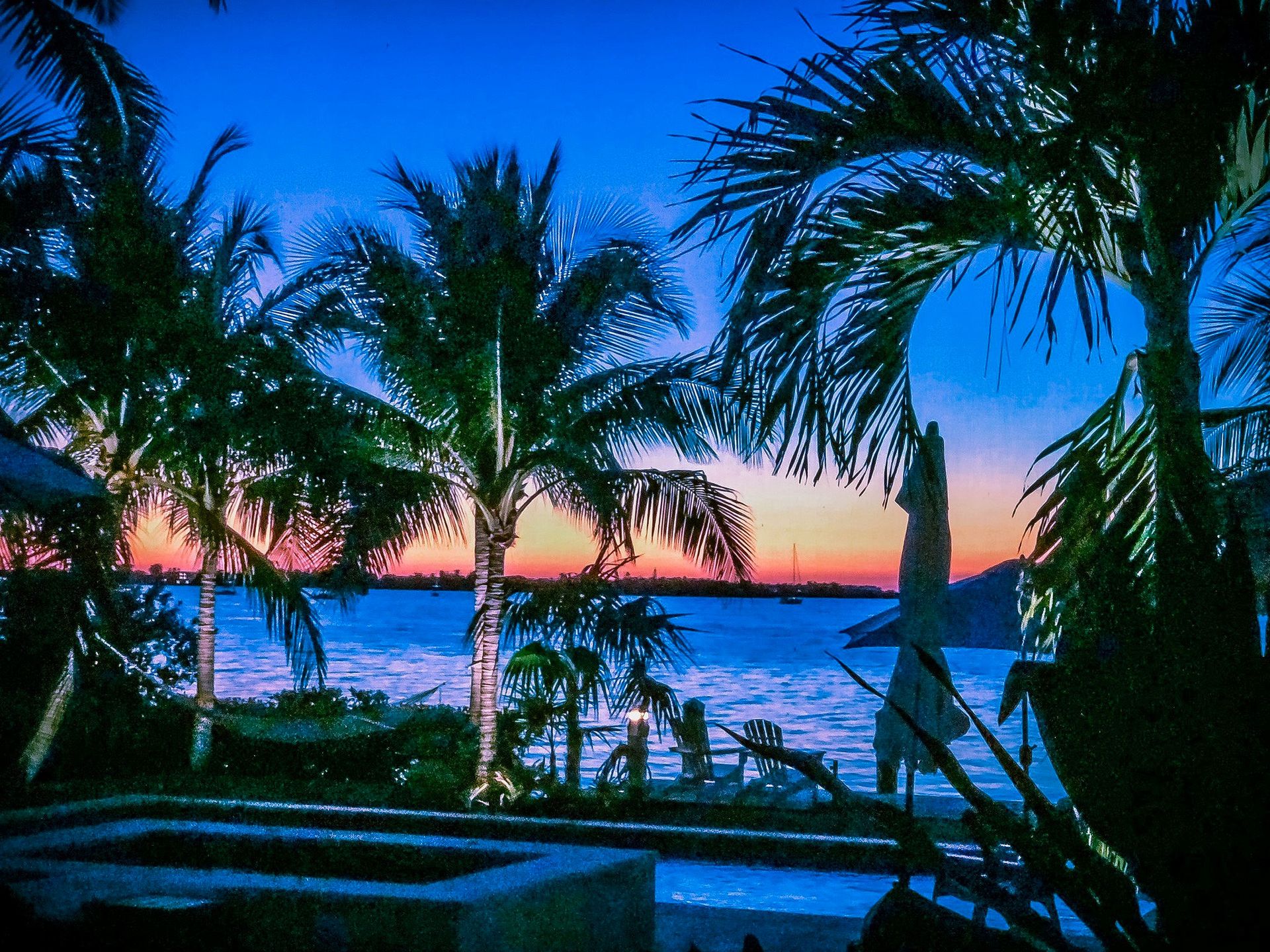 Palm trees silhouetted against a vibrant sunset over a calm, blue tropical bay near a pool deck.