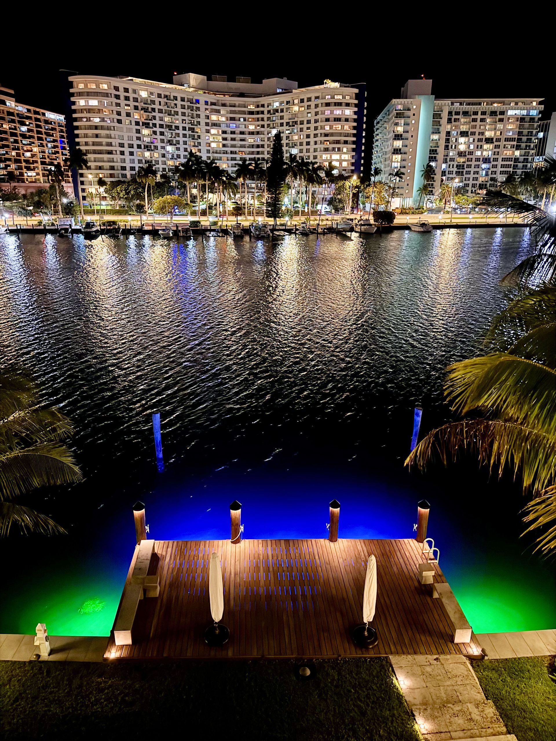 A nighttime view of a wooden dock with white umbrellas, overlooking a waterway with illuminated buildings in the distance.