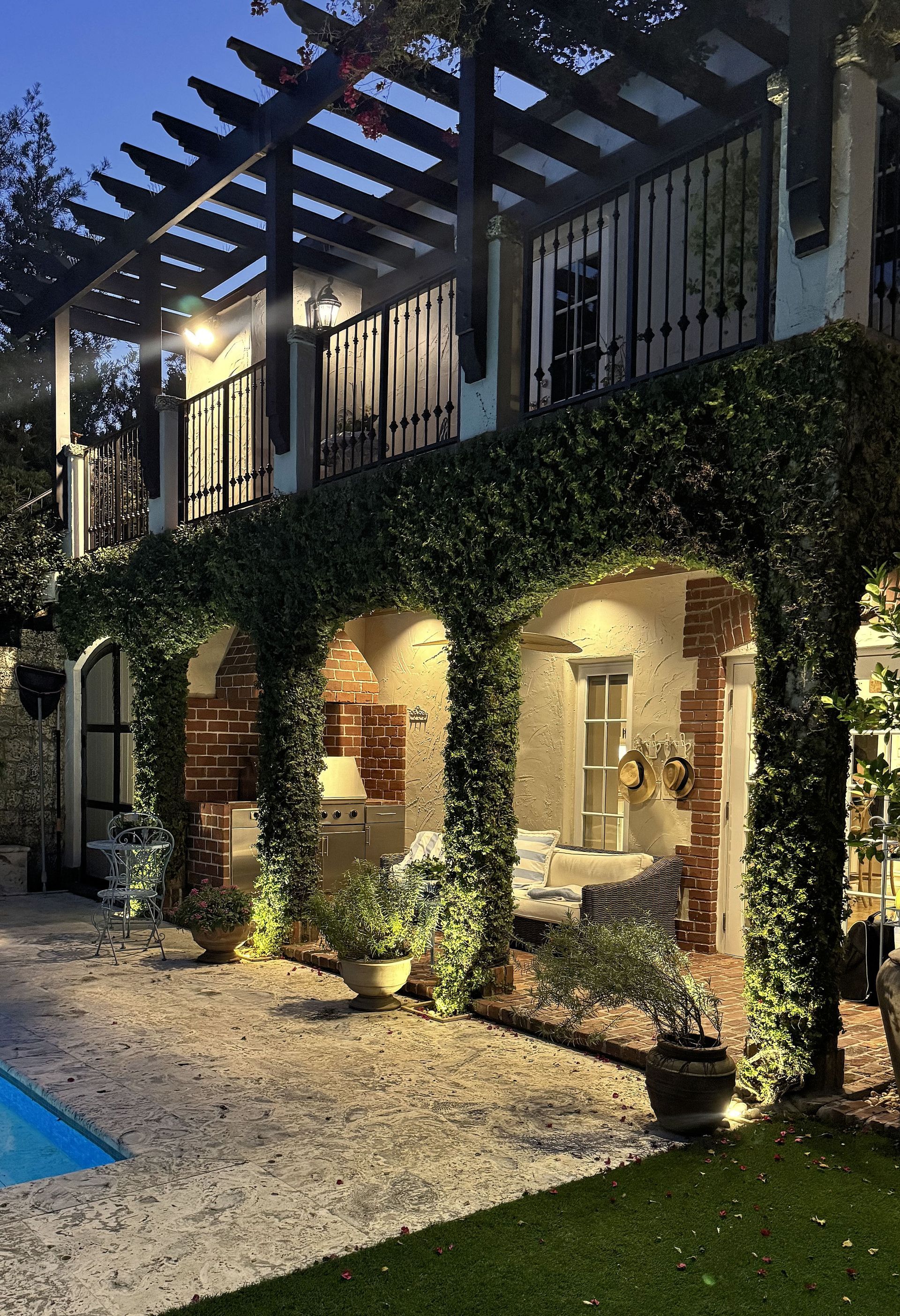A two-story villa with a vine-covered loggia, warm lighting, and a gravel patio next to a swimming pool at dusk.