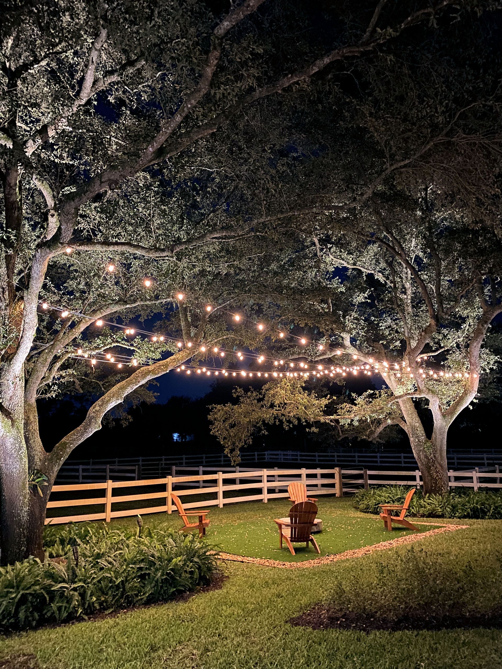 Nighttime garden scene with string lights hung between two large trees, three wooden chairs, and a white picket fence.