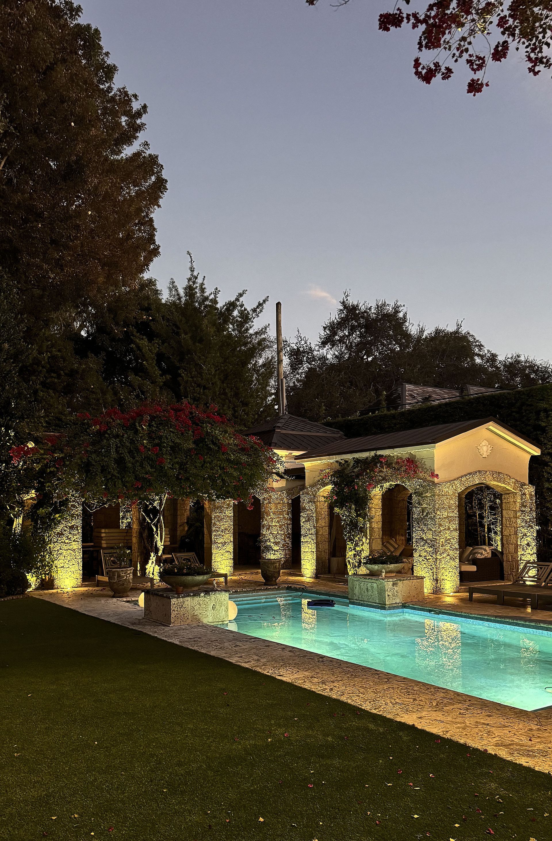 A lit outdoor swimming pool and stone patio at dusk, framed by lush trees and a stone structure with arched doorways.
