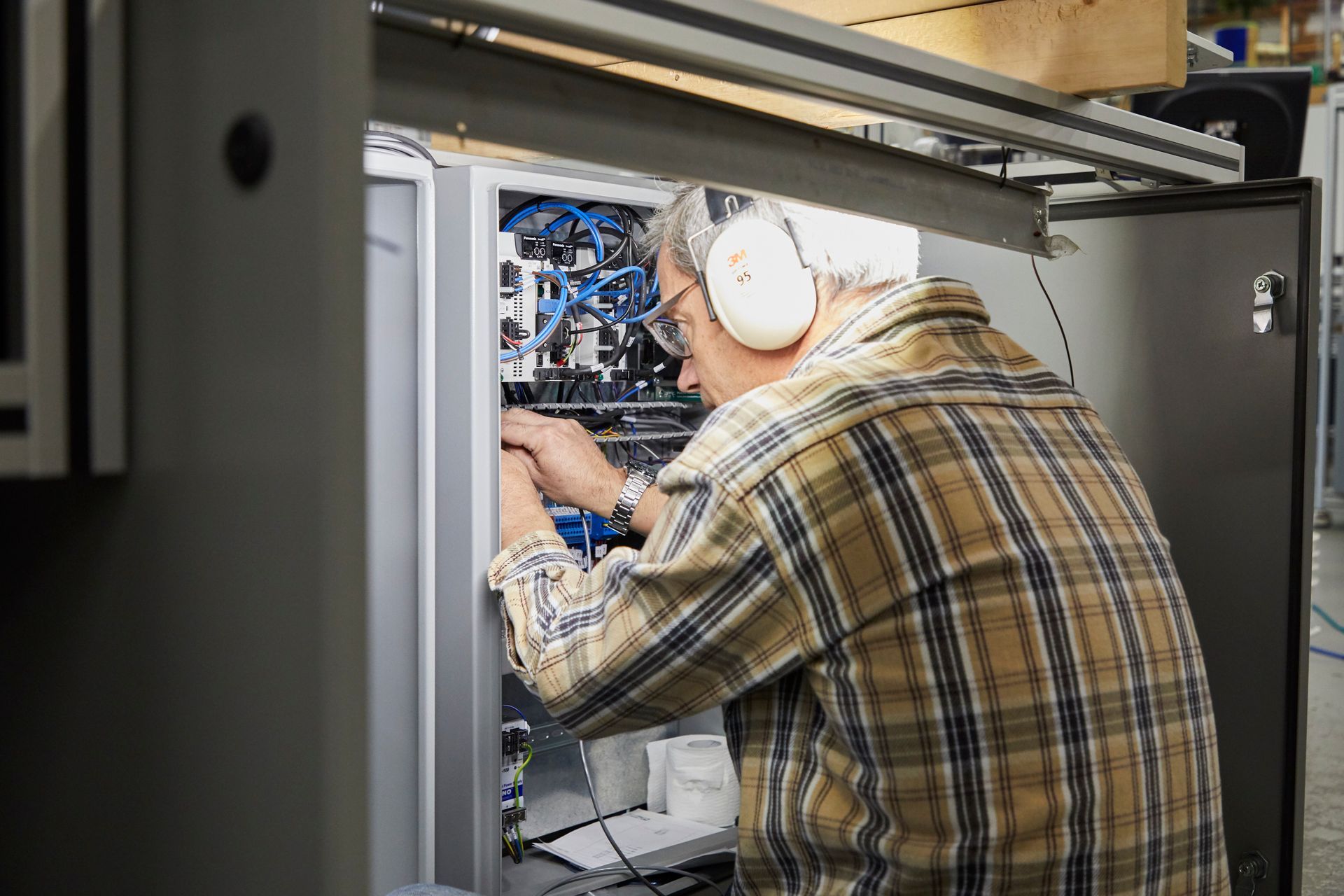 Technician in plaid shirt and headphones working inside an open server rack with blue wiring.