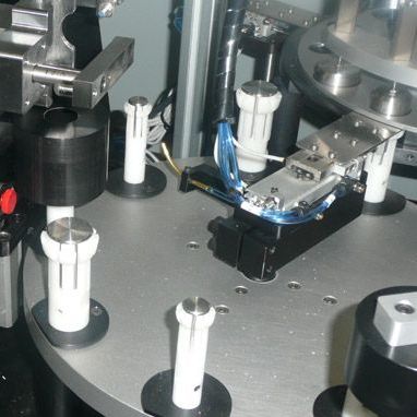 Metallic industrial machine with white posts, clamps, and blue wiring on a gray platform.