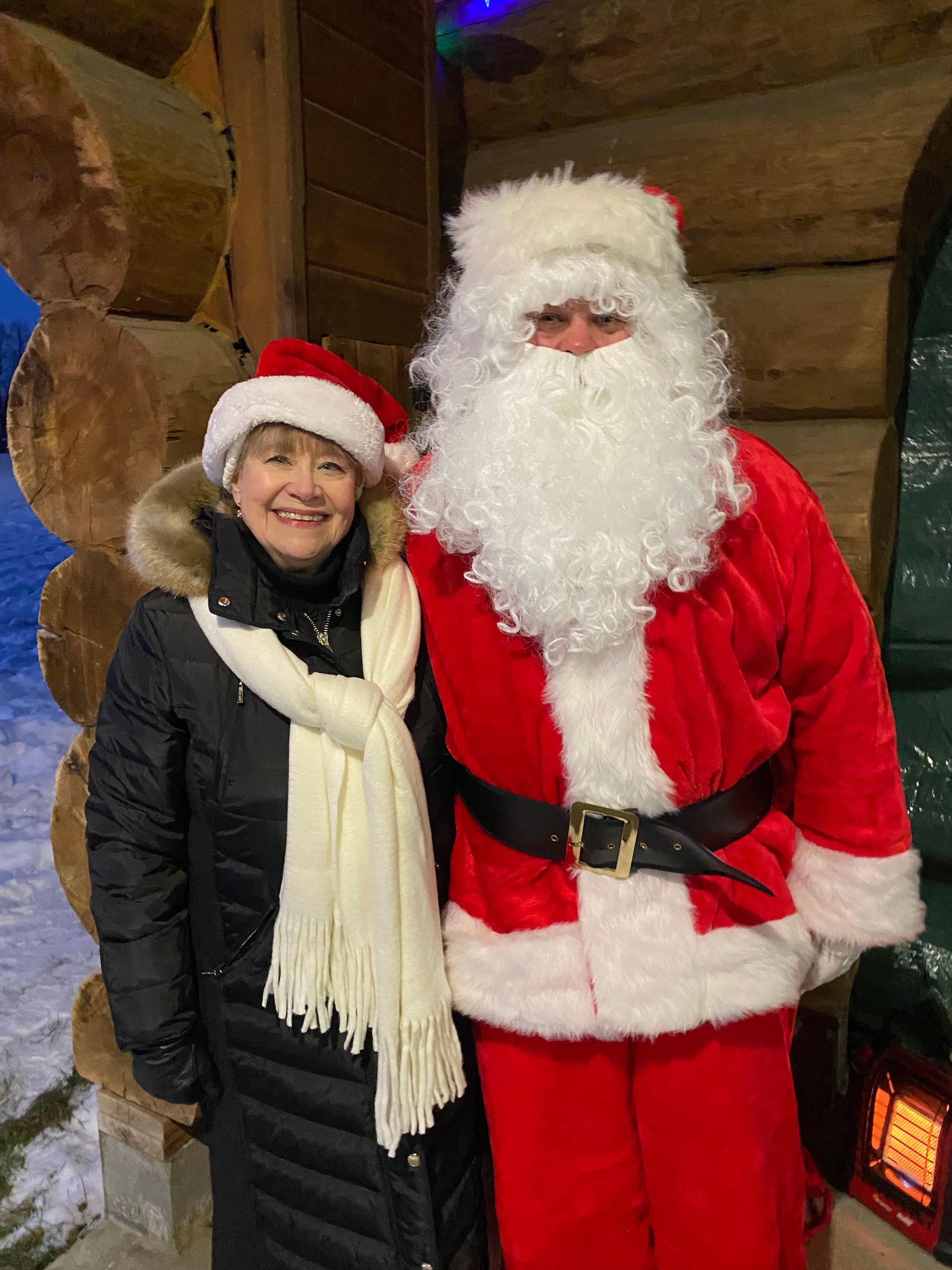 Santa Claus stands next to a smiling person in a winter coat, hat, and scarf inside a log cabin.