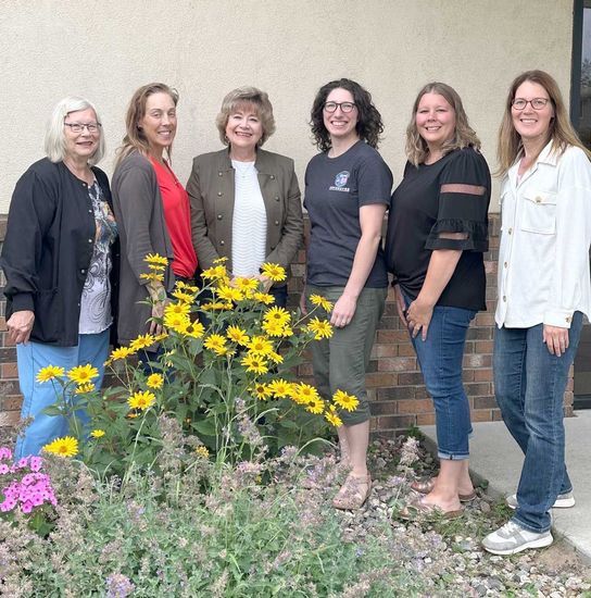 Six people stand in a line outdoors in front of a building, posed behind a flower bed with yellow sunflowers.