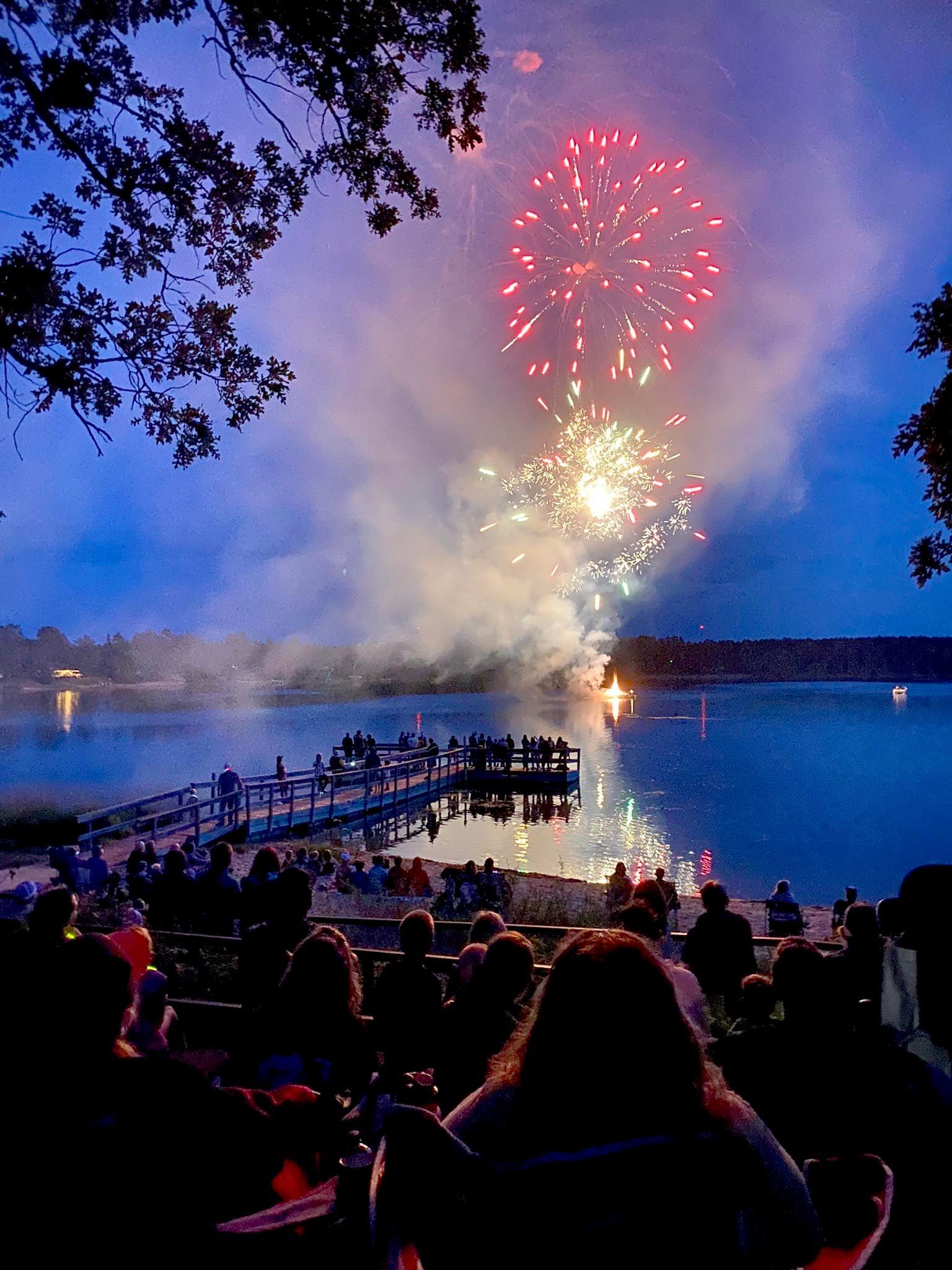Fireworks bursting over a lake at dusk, with a crowd watching from the shoreline and a dock.
