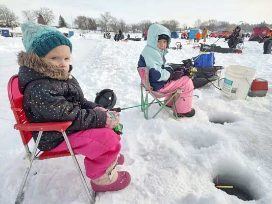 Two children in winter gear sit in folding chairs on a frozen, snow-covered lake, actively ice fishing near drilled holes.
