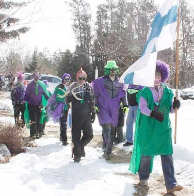 People in colorful costumes and wigs walk through a snowy area, with one person carrying a Finnish flag.