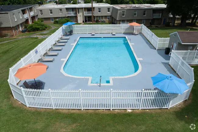 An aerial view of a swimming pool surrounded by a white fence, with umbrellas and buildings in the background at New Irving Heights, which offers apartments for rent in Greensboro, NC.