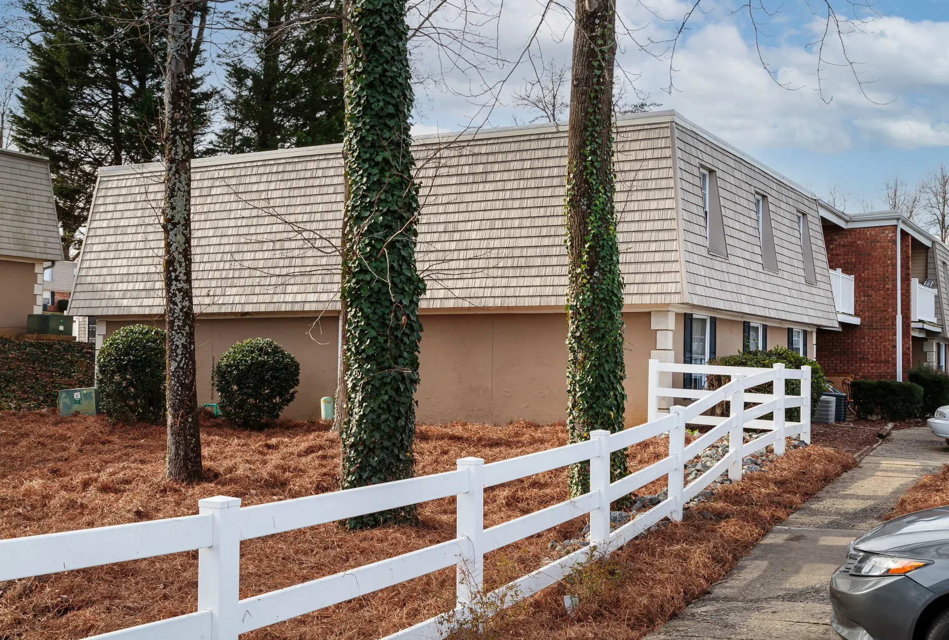 Exterior view of a beige apartment building with a white fence, ivy-wrapped trees, and trimmed bushes.