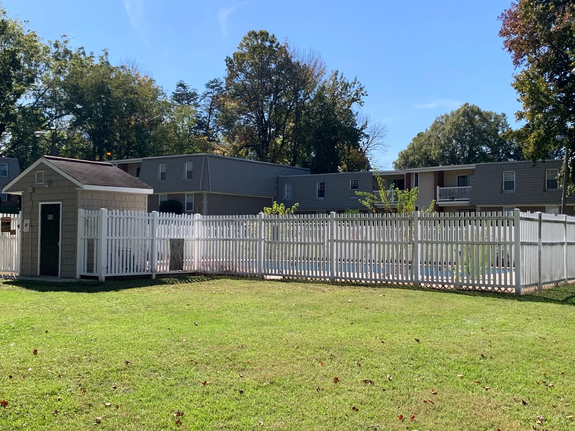 White fence and shed in grassy yard with mobile homes under a clear blue sky.