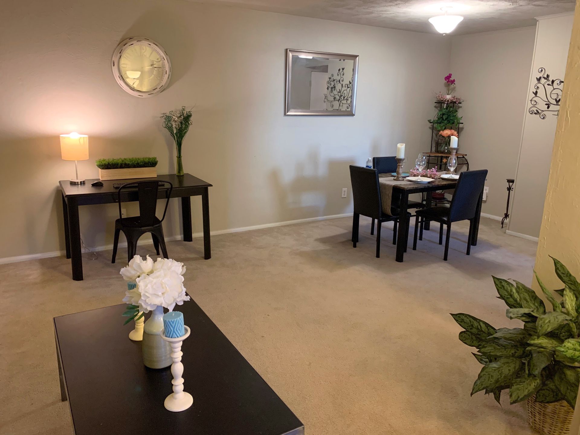 Spacious dining area with two small tables, chairs, a wall clock, and potted plants on carpeted floor.
