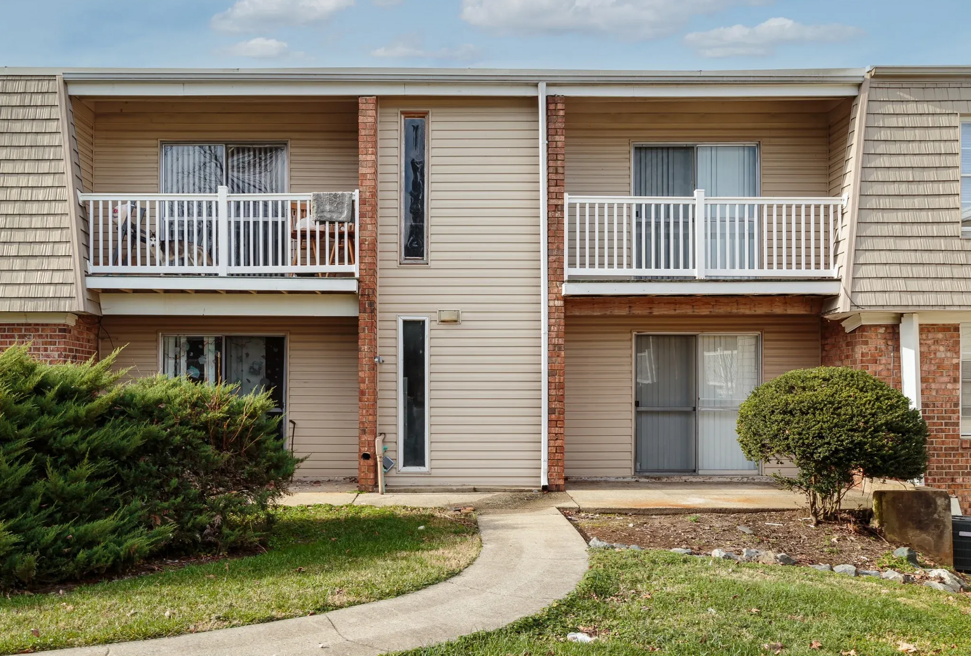 Exterior of a two-story apartment building with beige siding, brick columns, and white balcony rails.