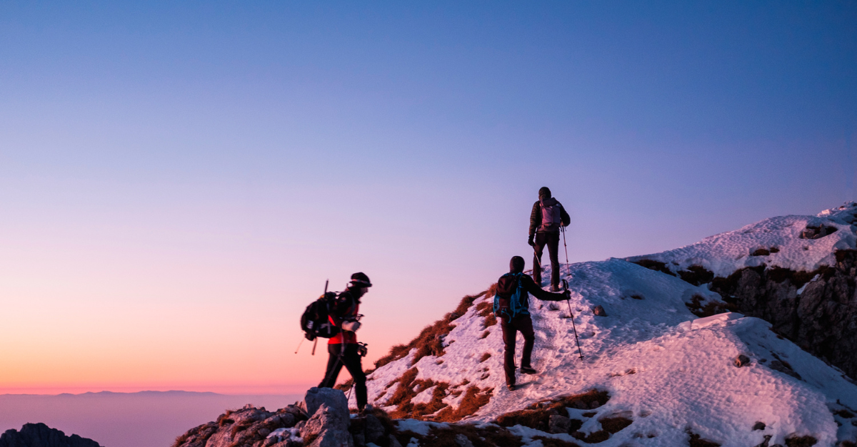 Three hikers climb a snowy mountain ridge at sunrise under a pink-blue sky