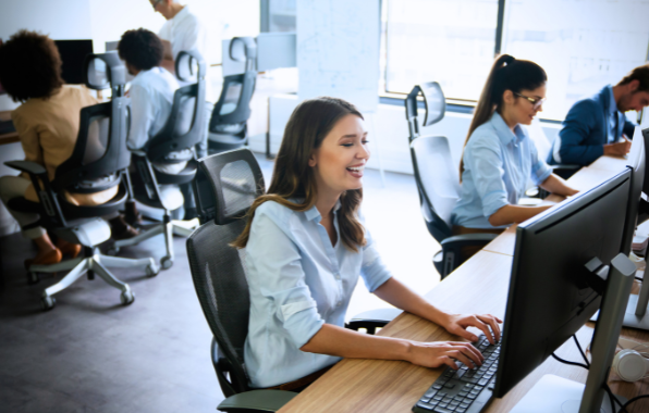 People working at desktop computers in a bright open office, with one woman typing in the foreground.