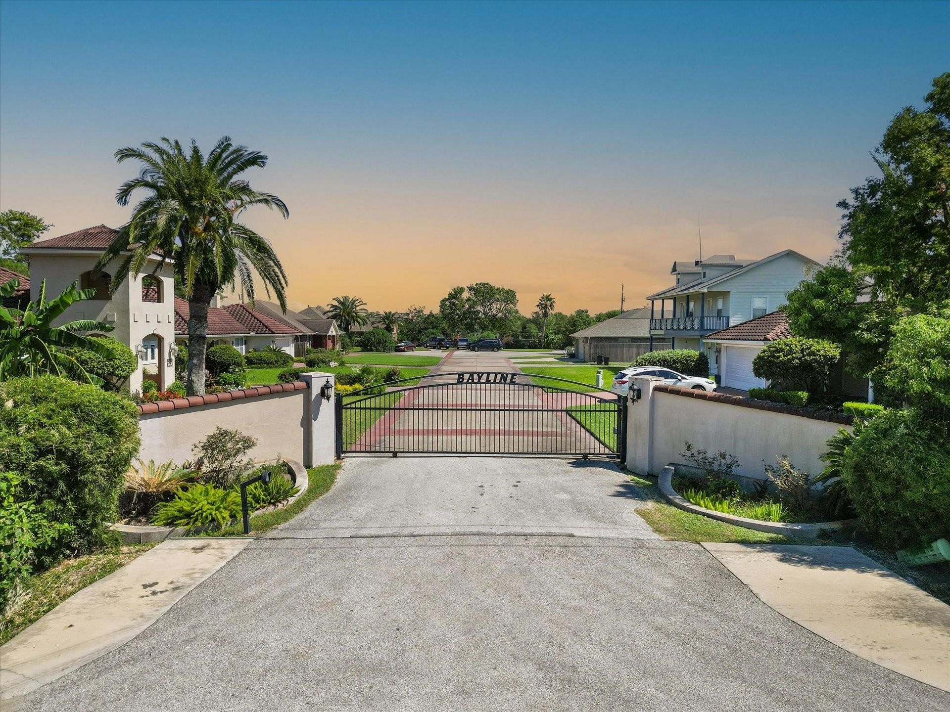 Gated community entrance with closed metal gate. Beige walls, palm tree, and houses visible.