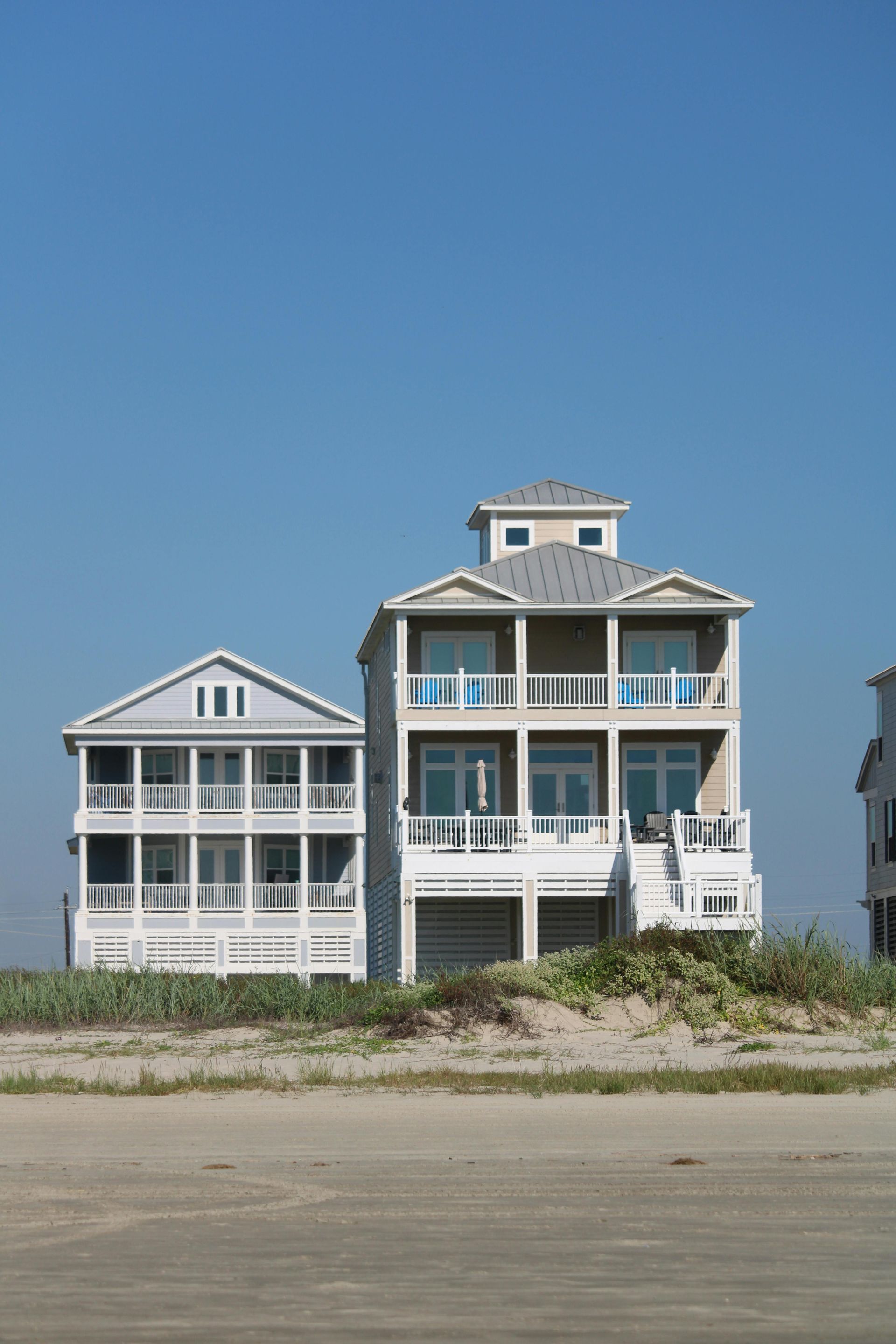 Two-story beach houses on a sandy shore under a clear blue sky.