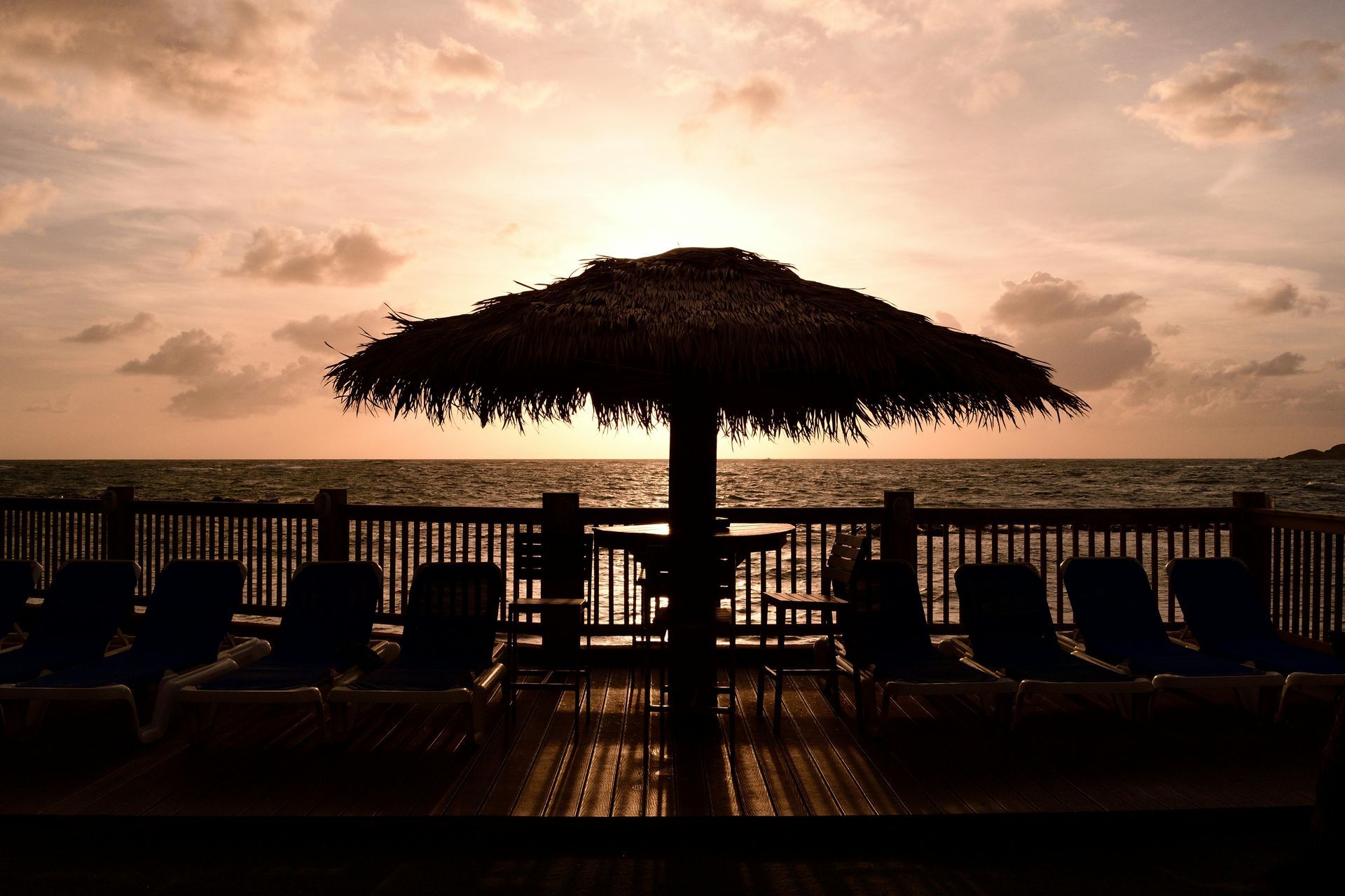 Silhouette of deck with umbrella, chairs, and ocean view at sunset.