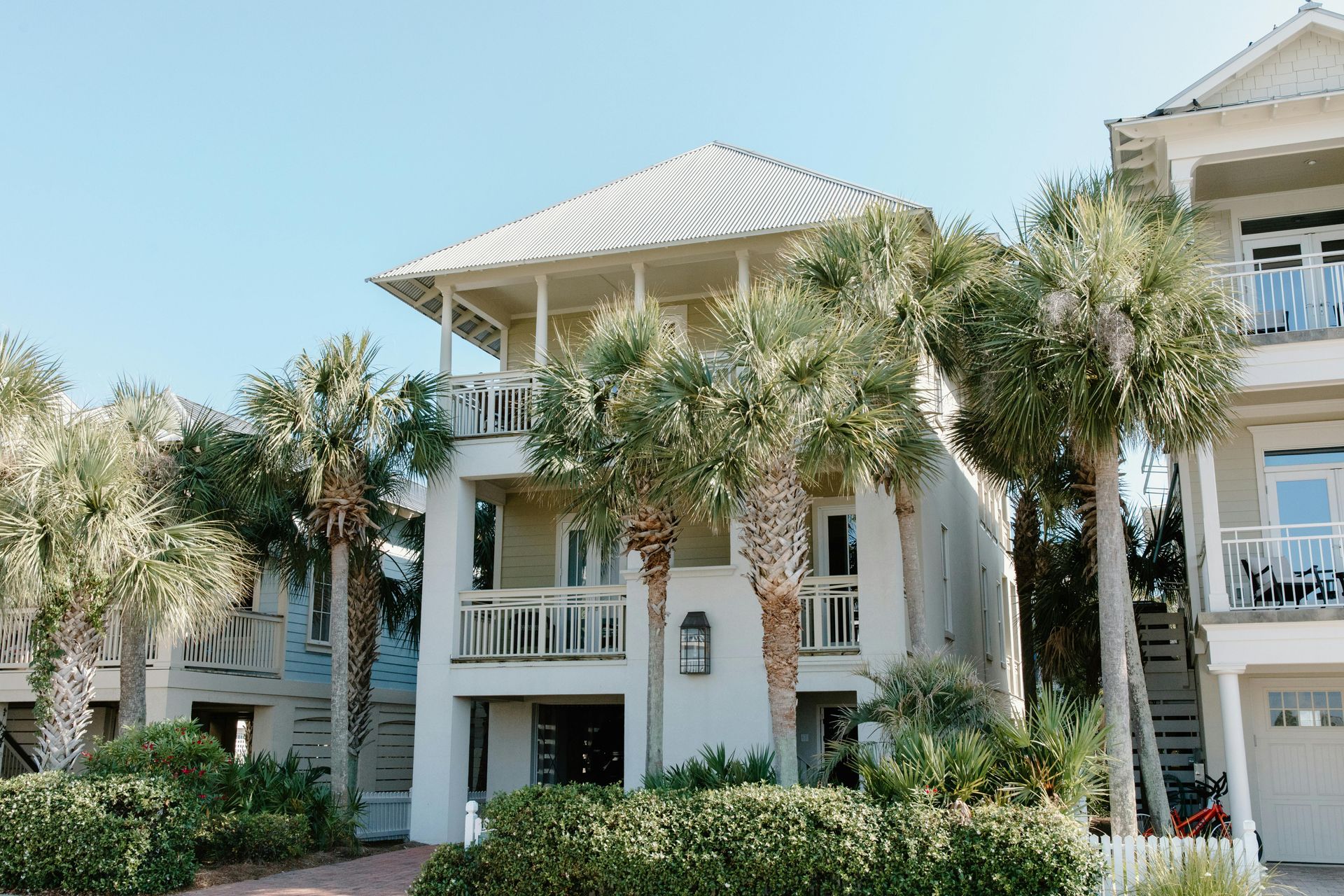 Three-story white beach house with balcony surrounded by palm trees under a blue sky.