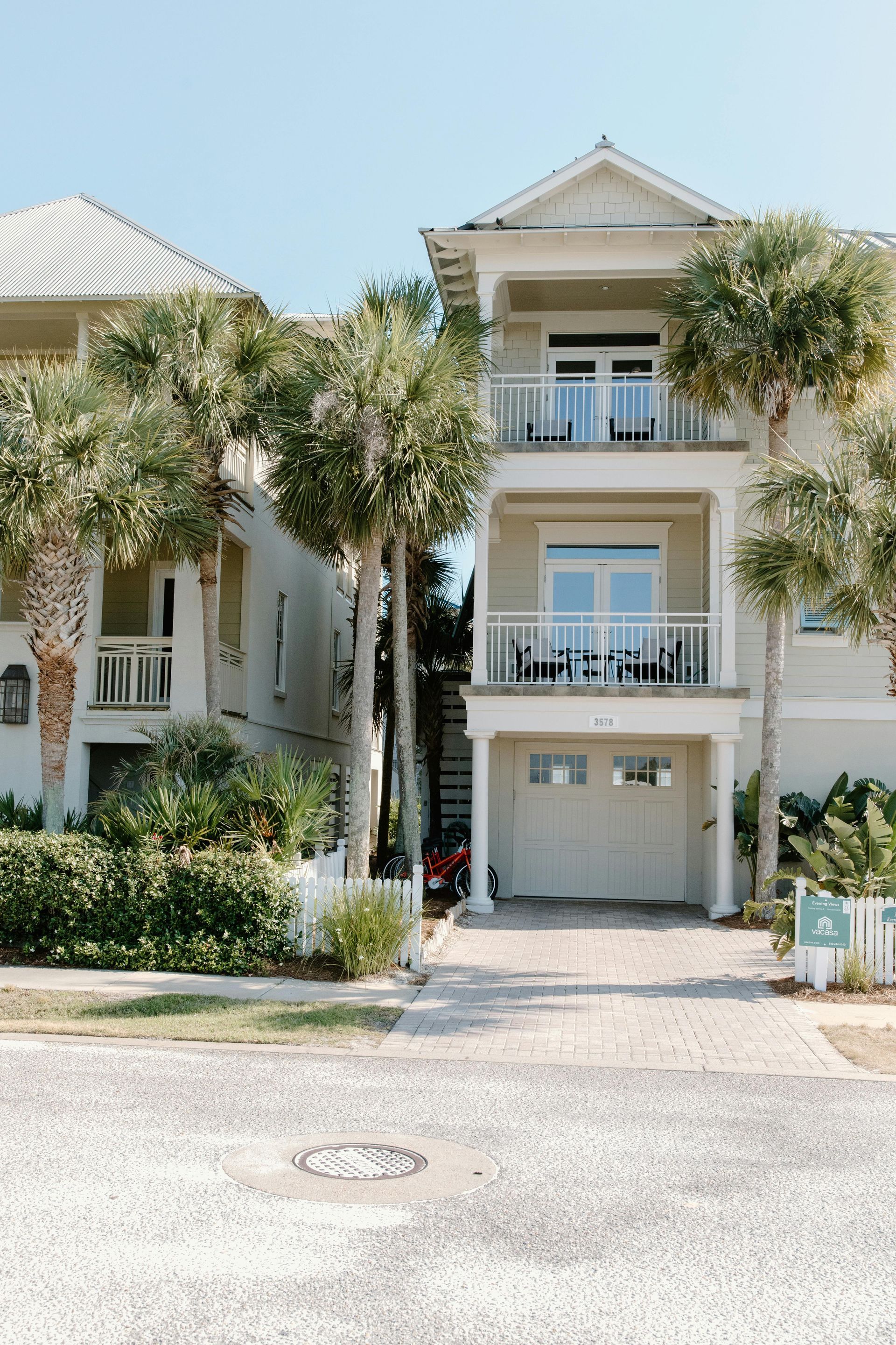 Multi-story beach house with balconies, palm trees, and driveway.