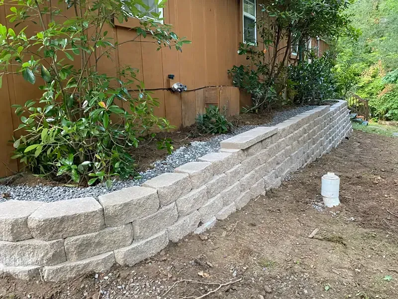 Stone retaining wall along the side of a brown house with shrubs and a dirt yard