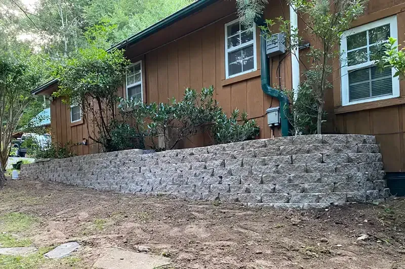 Wooden house with a gray stone retaining wall and garden plants along a sloped dirt yard.