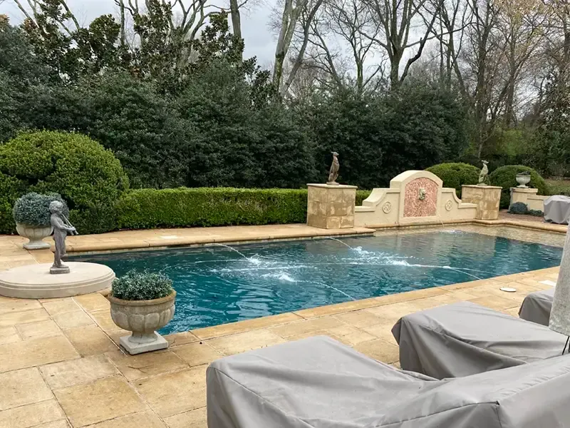 Backyard pool with tan stone deck, gray loungers, potted plants, and trimmed hedges