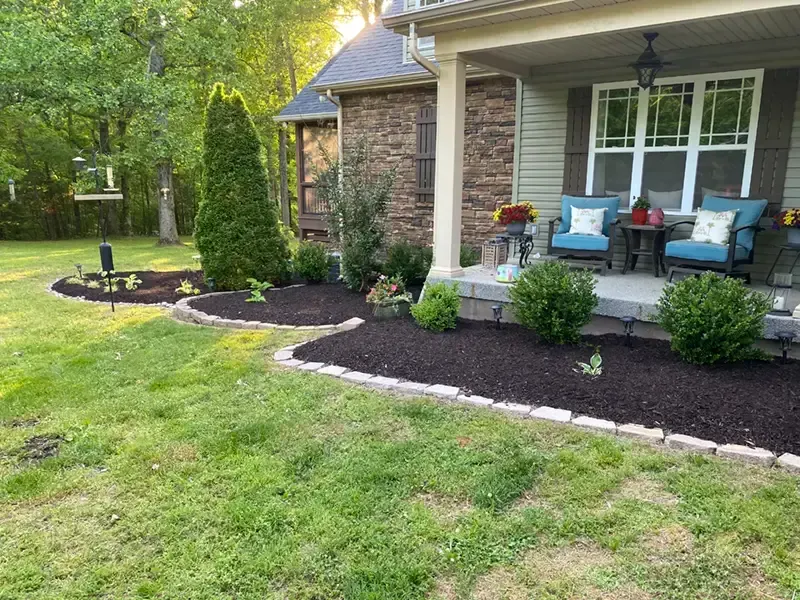 Front porch with blue chairs, stone columns, and landscaped garden beds along a grassy yard.