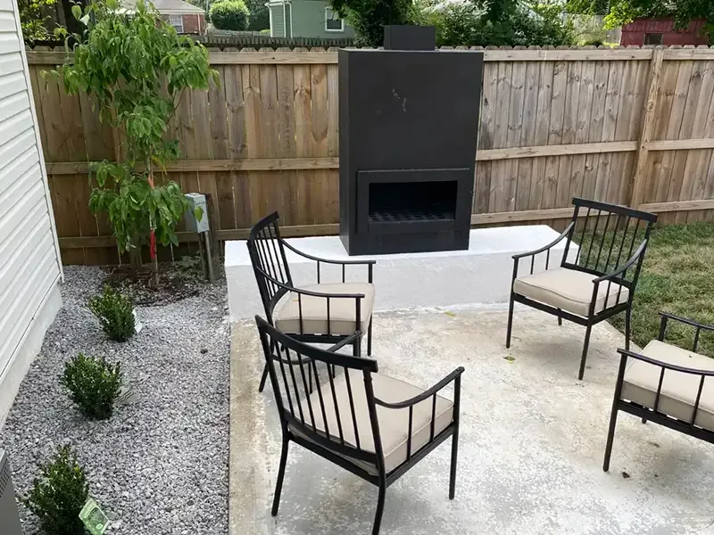 Patio seating area with four black chairs around a black outdoor fireplace on a gravel and concrete yard