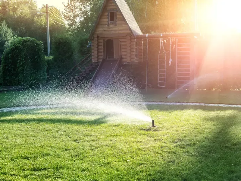 Lawn sprinkler spraying water in a sunlit backyard with a wooden shed and climbing frame