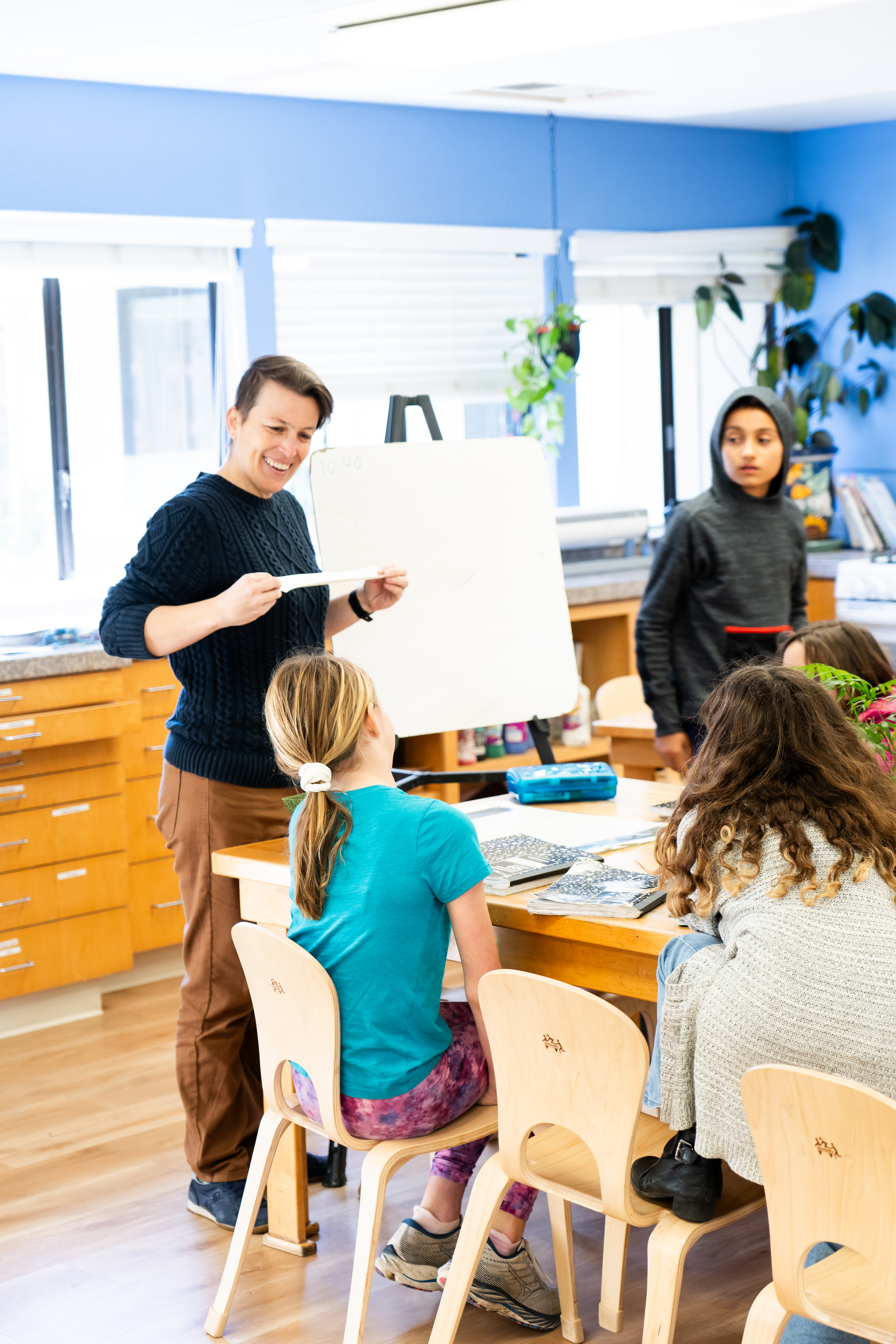 A Montessori guide is standing in front of a group of children sitting at a table.