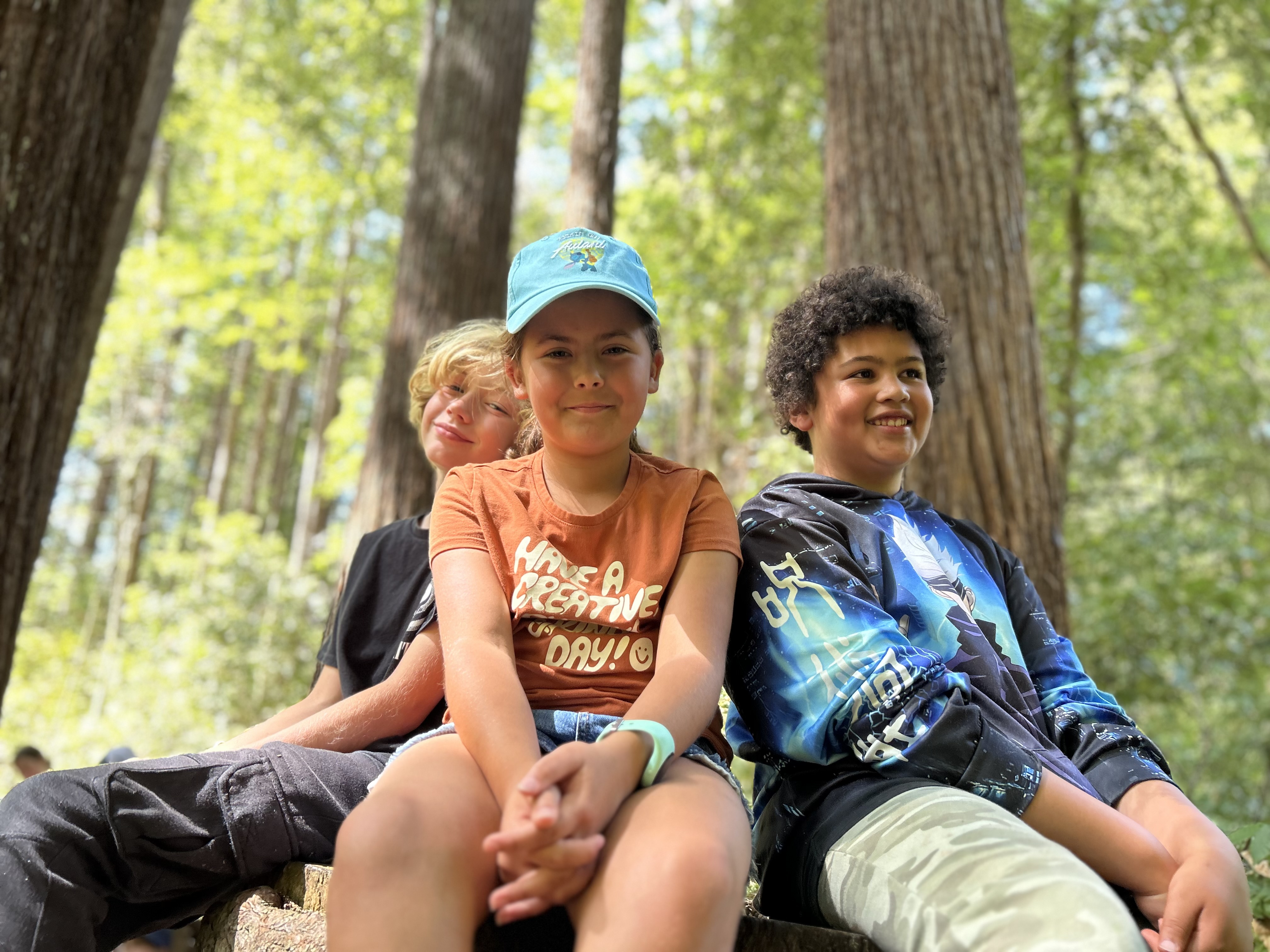 Three Montessori children are sitting on a log in the woods.