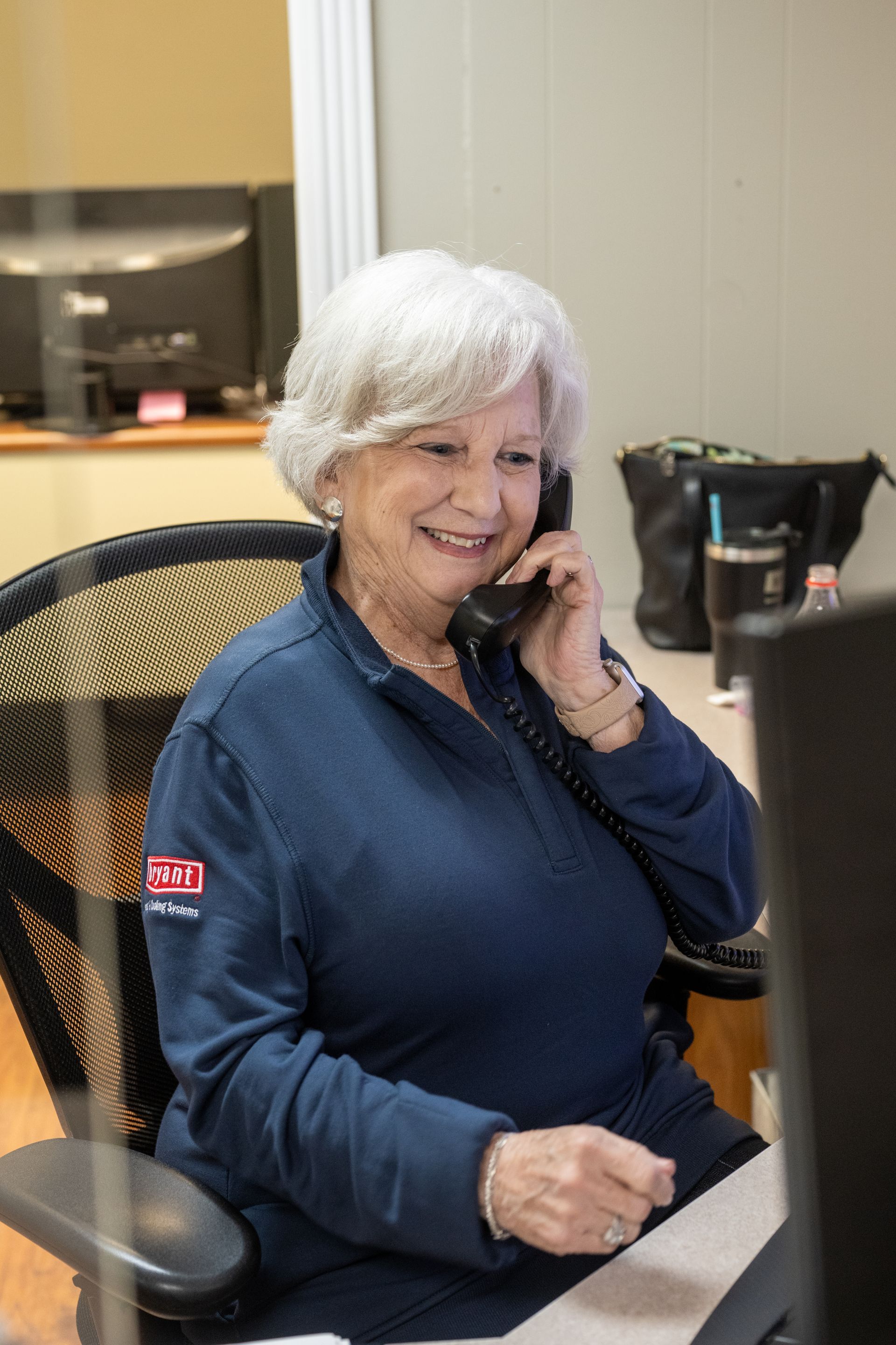 A person in a blue long-sleeved shirt sitting at a desk, smiling while talking on a landline telephone in an office.