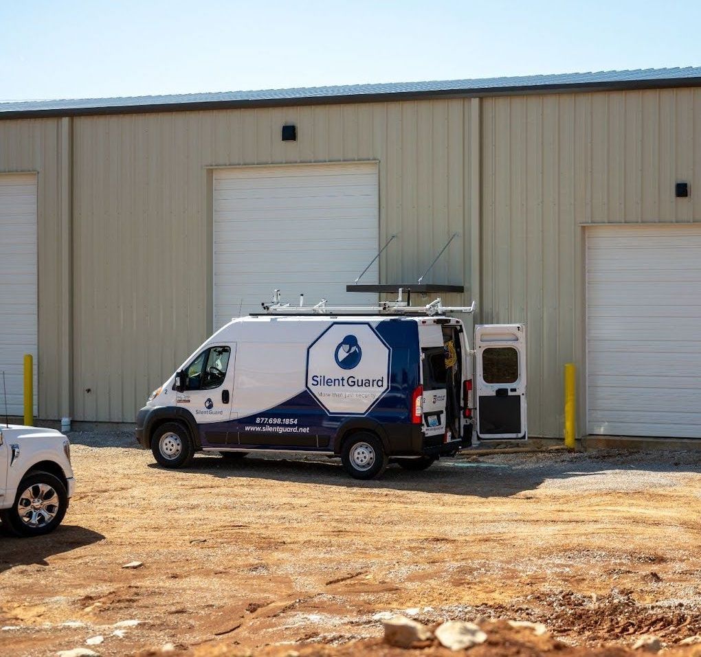 A white utility van with a blue logo parked in front of a metal warehouse building on a dirt lot.