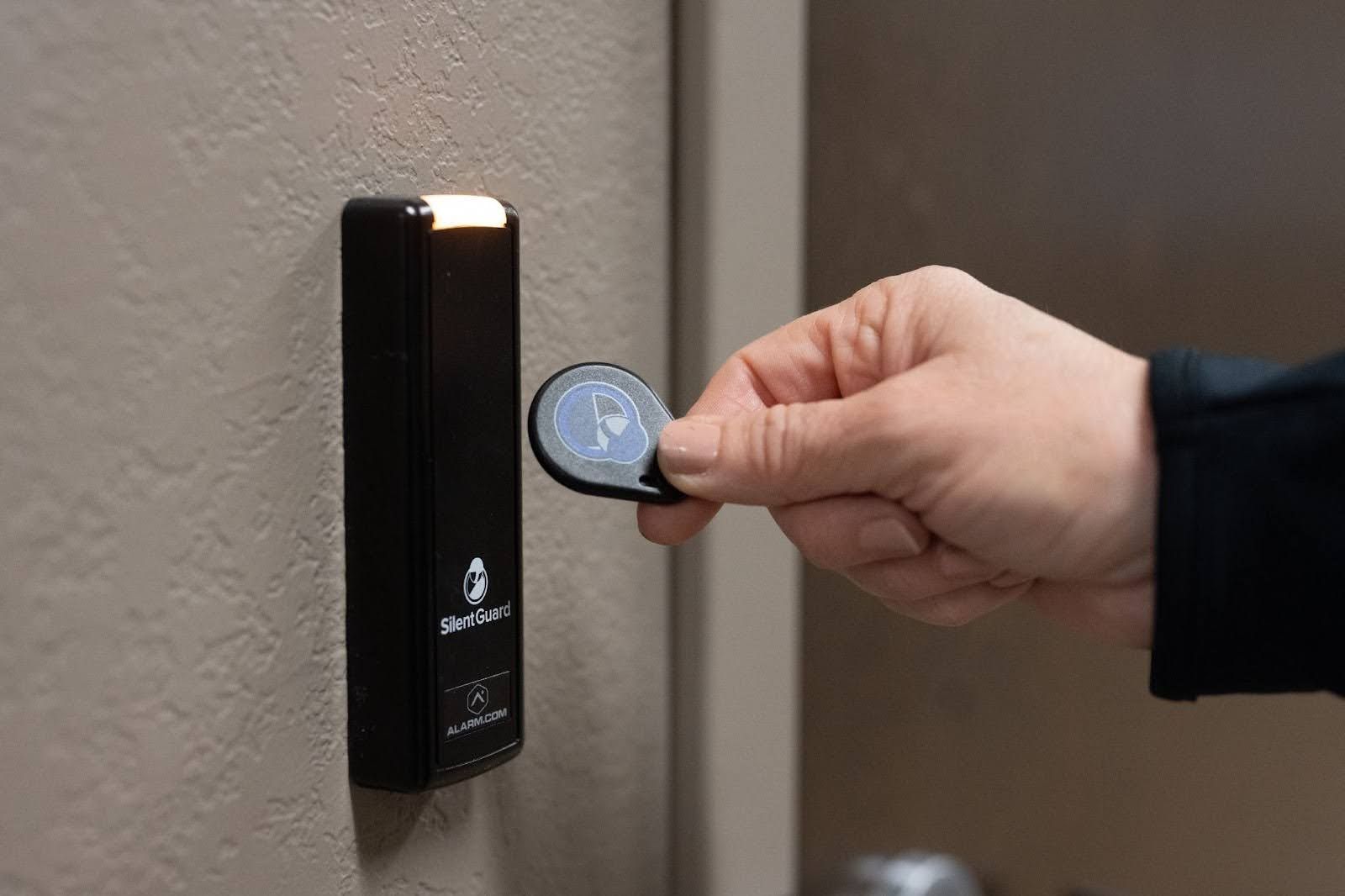 A hand holds a black key fob near a wall-mounted electronic access control reader with a glowing light.