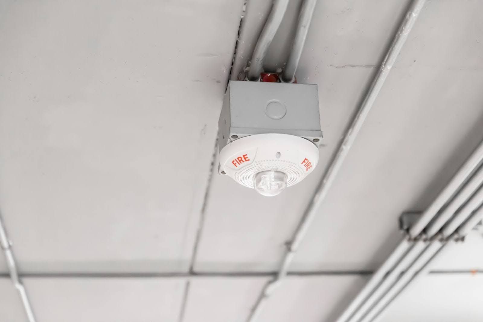 A white circular smoke or heat detector mounted on a gray junction box attached to a light-colored concrete ceiling.