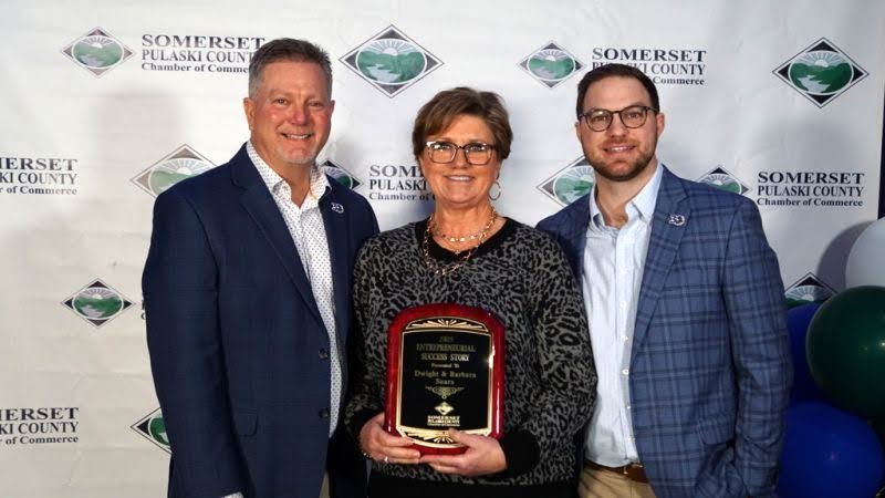 Three people stand smiling in front of a Somerset-Pulaski County Chamber of Commerce banner, holding an award plaque.