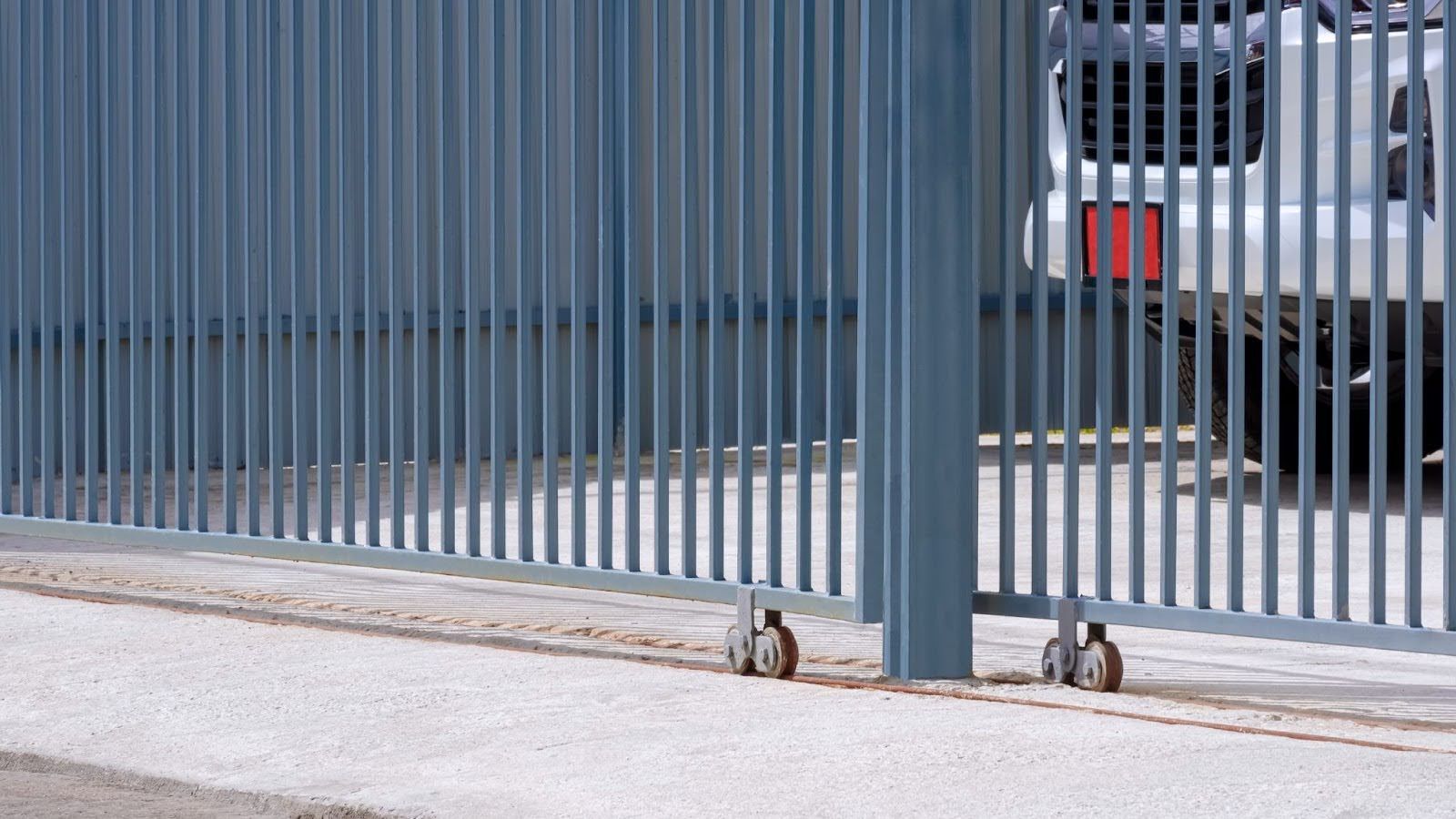 Gray metal sliding gate on tracks with small wheels, featuring vertical bars and a partial view of a white vehicle behind.