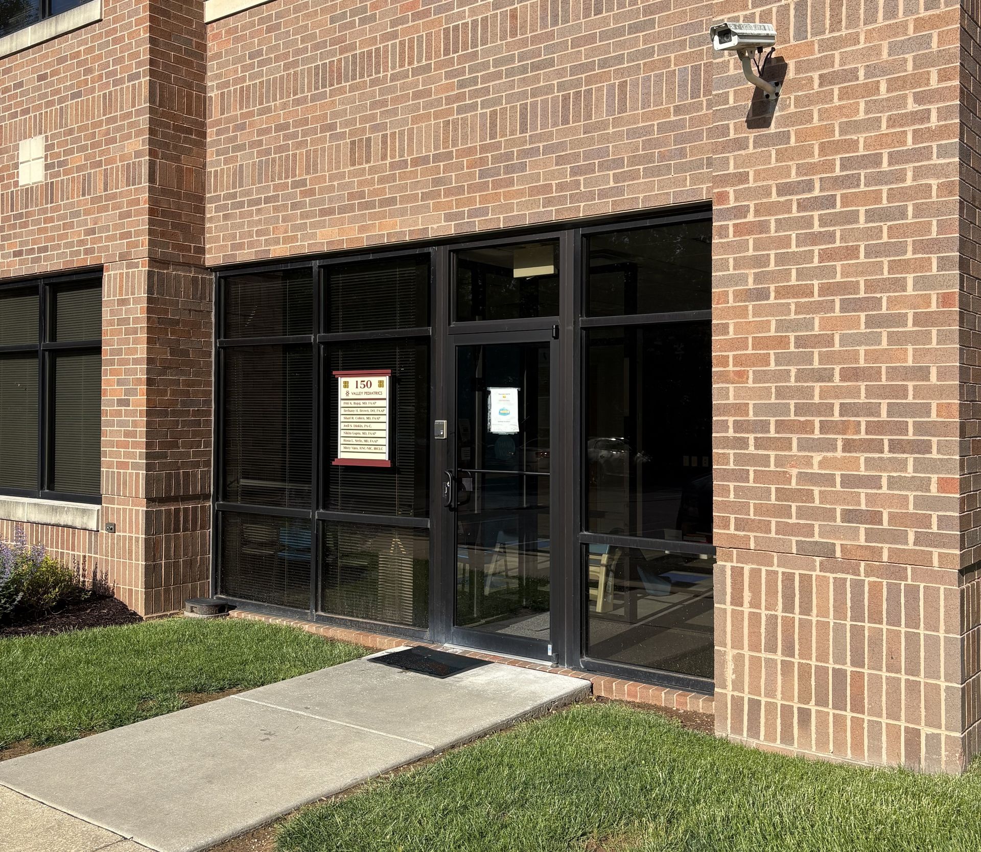 Brick building entrance with black-framed glass doors and windows. Concrete pathway and green grass.