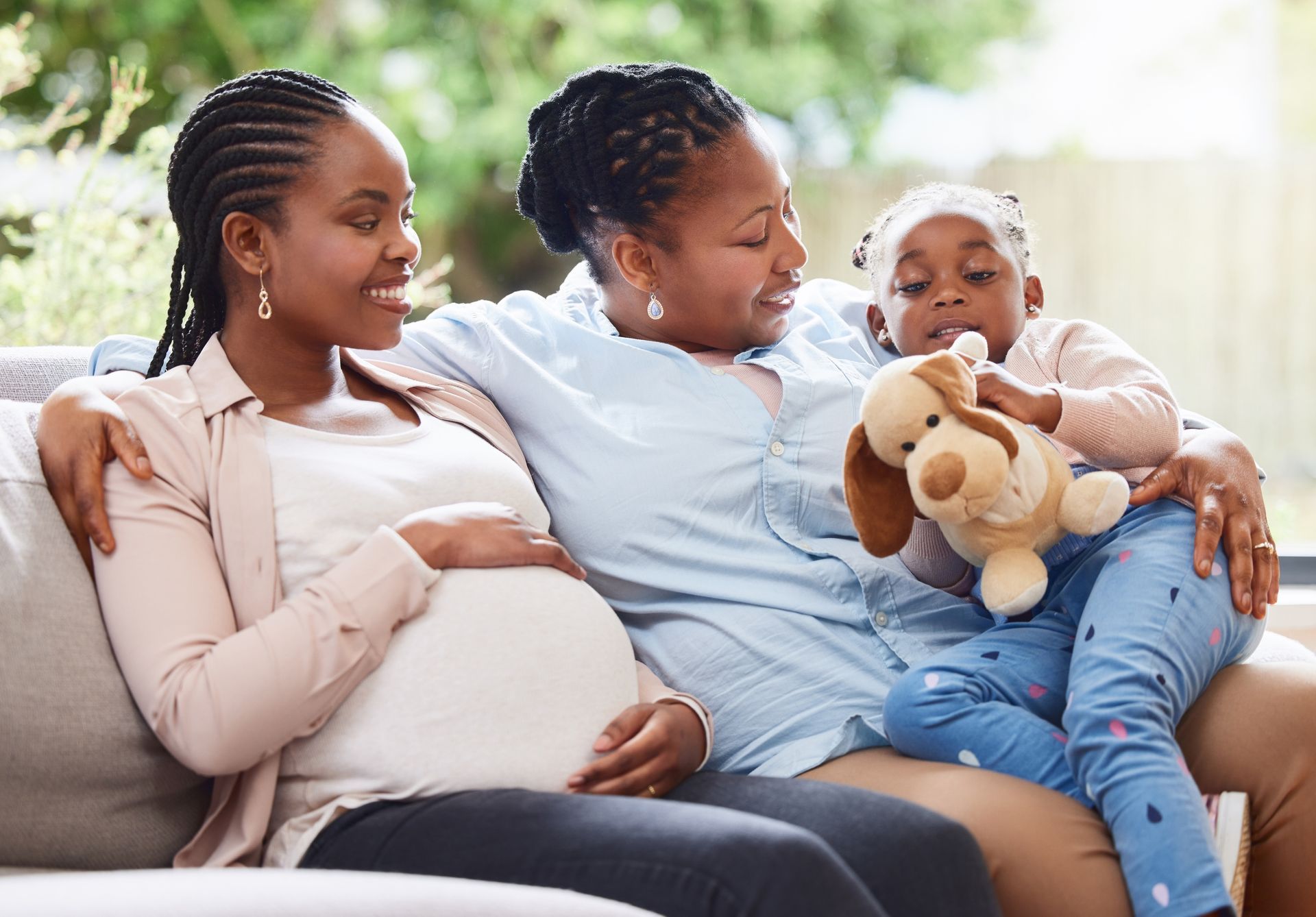 A pregnant woman, her mother and her daughter are all sitting on a sofa in a bright room.