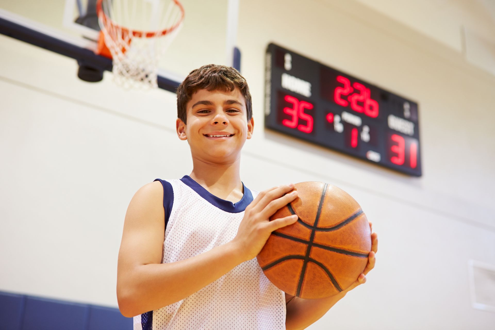 A boy is in a gym holding a basketball with a time clock and basketball hoop in the background.