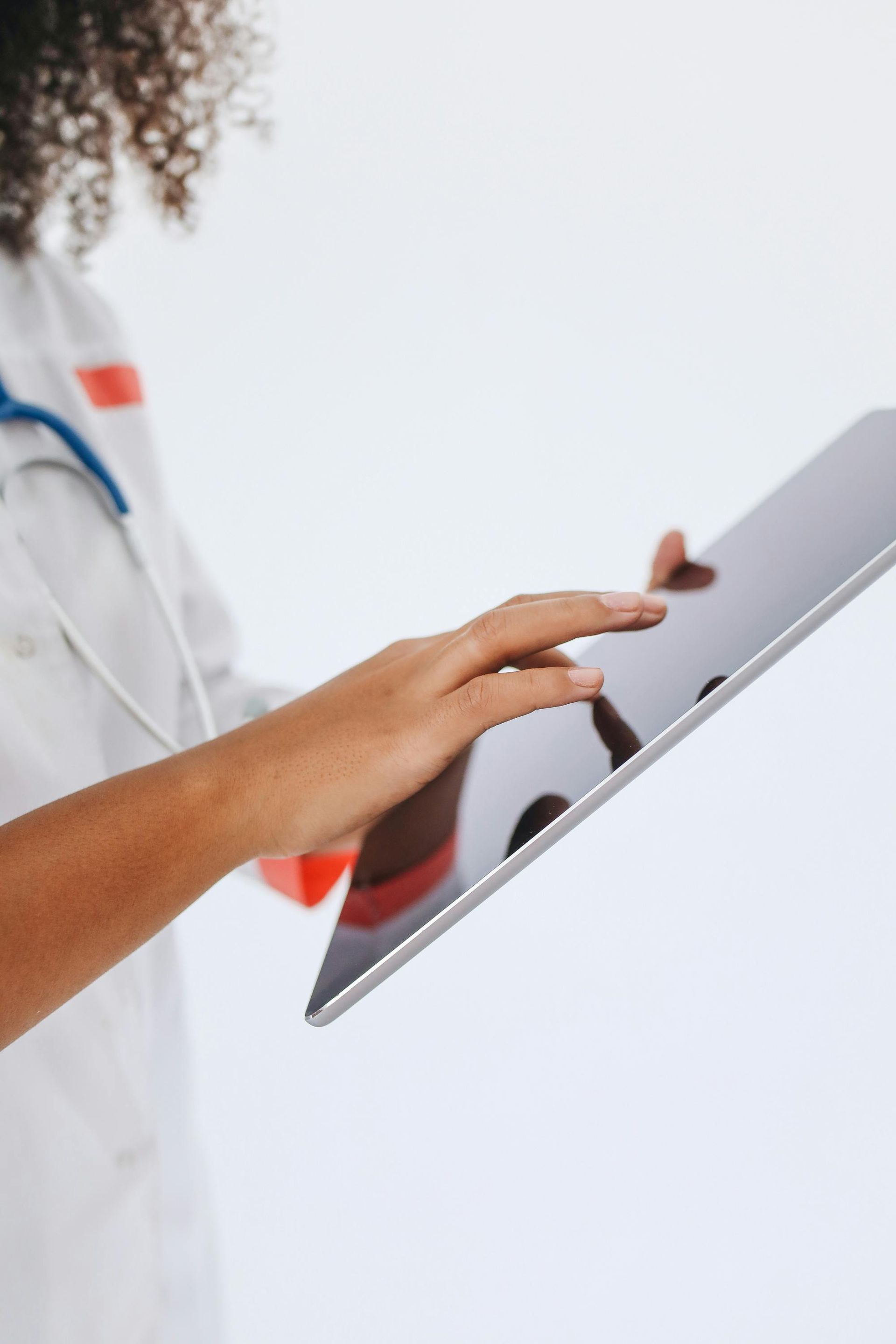 Doctor using a tablet; white coat, stethoscope, red name tag, light background.