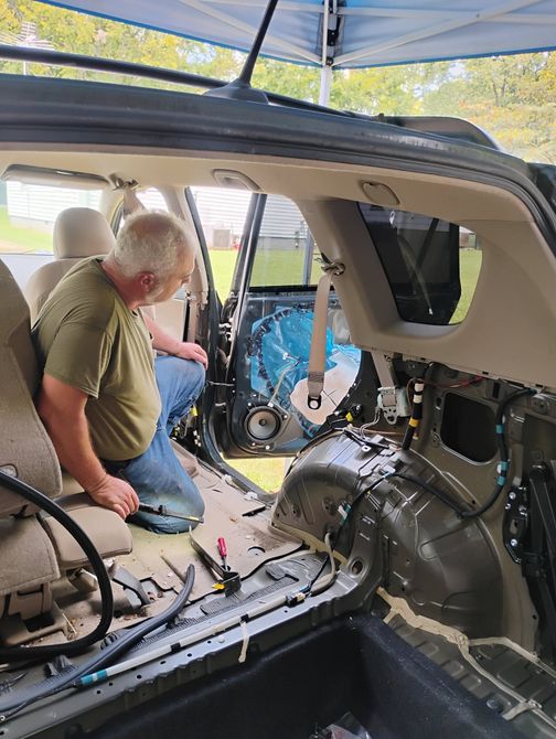 David Lutrick kneeling beside an open car hood in a customer driveway with tools laid out on a fender mat