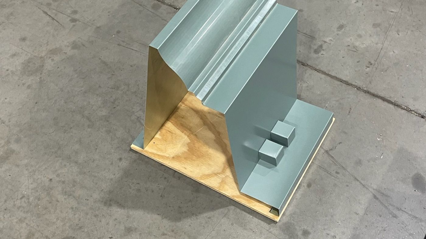 Green-gray formed assembly with curved and stepped visible geometry resting on a wooden base.