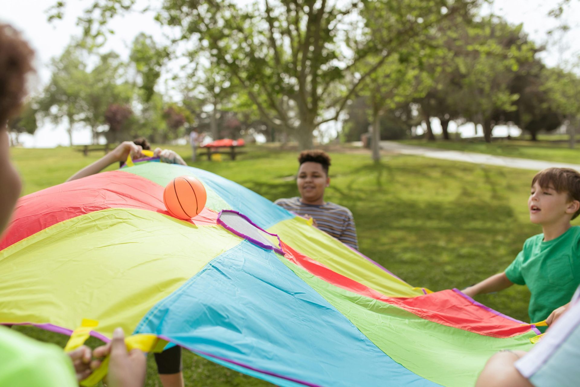 A group of children plays with a colorful parachute and a small ball in a park.