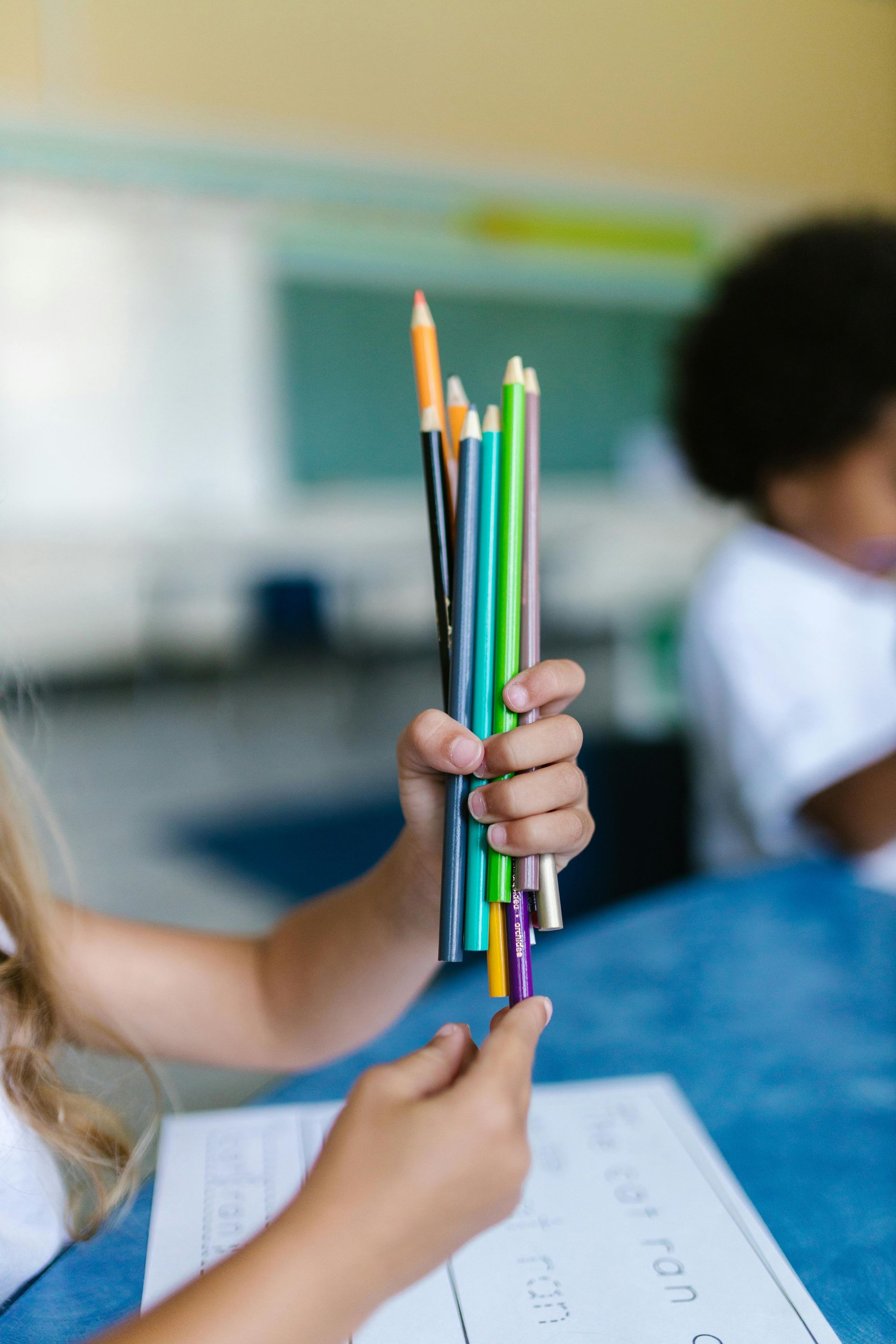 A person holds a bundle of multi-colored pencils over a worksheet in a classroom setting.