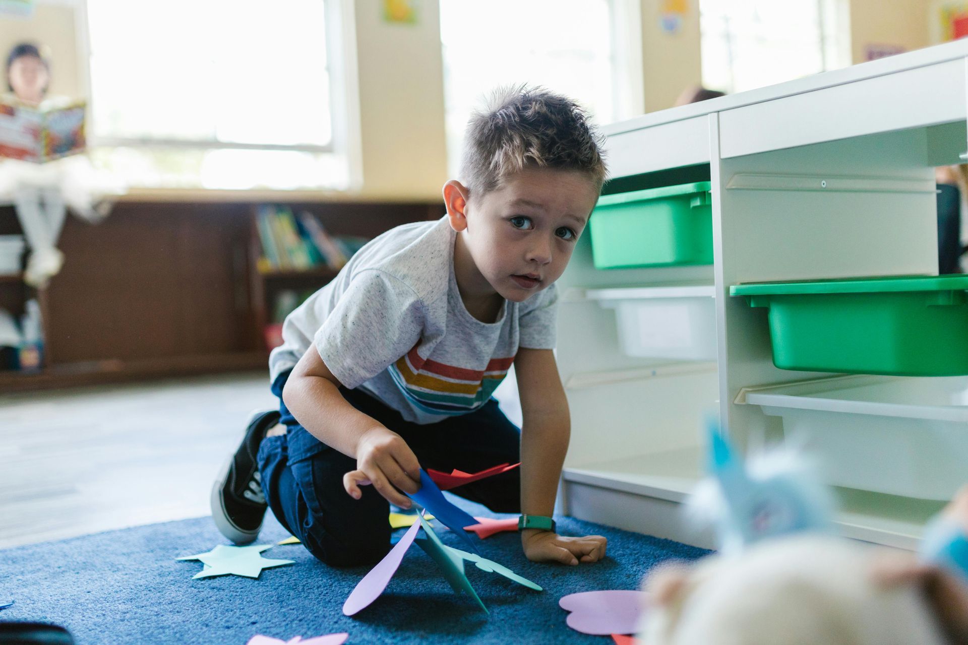 A child kneels on a blue rug, arranging colorful paper shapes in a bright classroom with storage cubbies.
